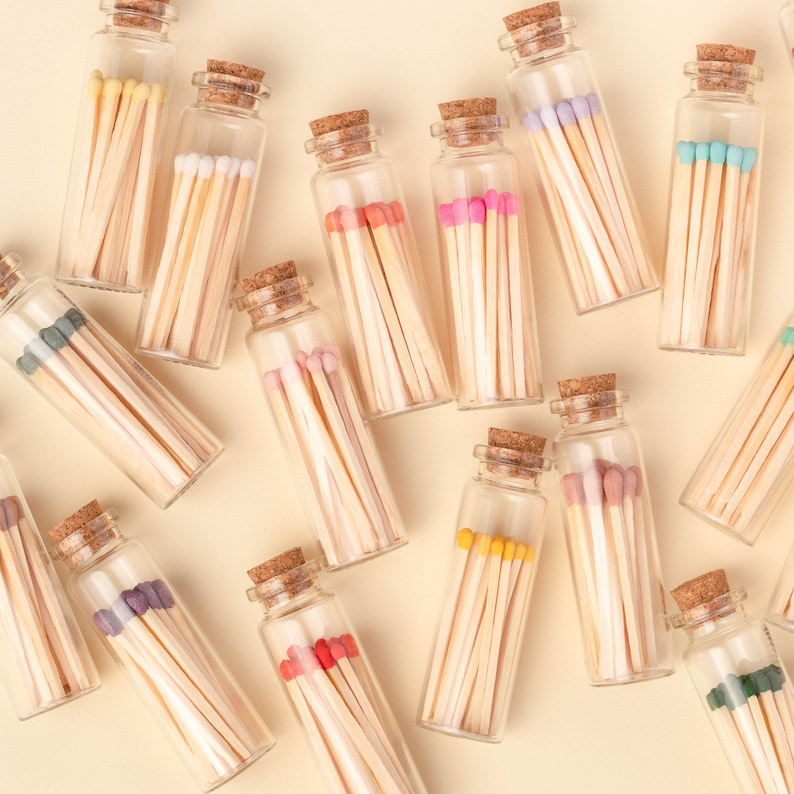 May include: Assortment of small glass bottles filled with colorful matchsticks. The bottles have cork tops and are arranged on a light beige background.