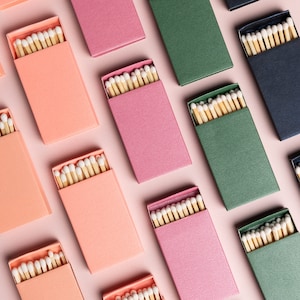 May include: A flat lay of various matchboxes with different colored lids and white matchsticks inside. The matchboxes are arranged in a grid pattern on a pink background.