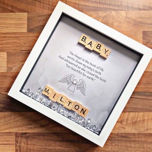 May include: A white framed artwork featuring the name "MILTON" in Scrabble tiles. The artwork also includes the word "BABY" in Scrabble tiles, an angel illustration, and a quote about a baby's birth. Clear crystals are scattered at the bottom.