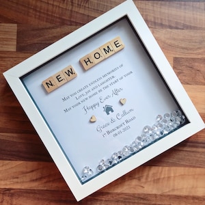 Pu&ograve; includere: Una cornice bianca con uno sfondo blu chiaro e le parole "NEW HOME" scritte con tessere di lettere in legno. Il testo recita "May you create endless memories of love, joy and laughter. May your new home be the start of your Happy Ever After." con i nomi "Grace & Callum", l'indirizzo "17 Burcroft Road" e la data "08-01-2021". La cornice &egrave; decorata con piccoli strass trasparenti.