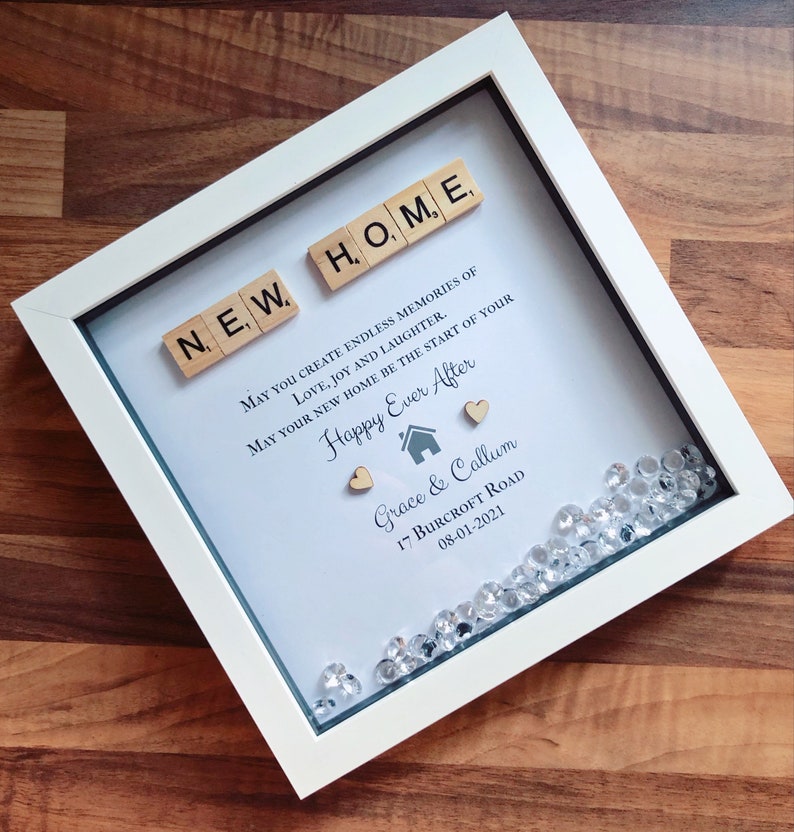 Pu&ograve; includere: Una cornice bianca con uno sfondo blu chiaro e le parole "NEW HOME" scritte con tessere di lettere in legno. Il testo recita "May you create endless memories of love, joy and laughter. May your new home be the start of your Happy Ever After." con i nomi "Grace & Callum", l'indirizzo "17 Burcroft Road" e la data "08-01-2021". La cornice &egrave; decorata con piccoli strass trasparenti.