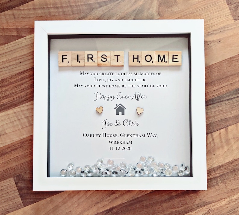Pu&ograve; includere: Una cornice bianca con un passepartout nero e uno sfondo bianco. La cornice contiene un messaggio che recita "FIRST HOME" scritto con tessere di lettere in legno. Il messaggio continua con "May you create endless memories of love, joy and laughter. May your first home be the start of your Happy Ever After." Il messaggio &egrave; seguito da una piccola icona di casa, due cuori e i nomi "Joe & Chris". I nomi sono seguiti dall'indirizzo "Oakley House, Glentham Way, Wrexham" e dalla data "11-12-2020". La parte inferiore della cornice &egrave; piena di gemme scintillanti e trasparenti.