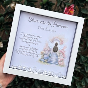 May include: A framed memorial artwork with the text "Staircase to Heaven" and "Our Lauren." The image depicts a staircase leading to an archway, with angel wings and a pebble figure. The white frame is decorated with clear crystals along the bottom edge.