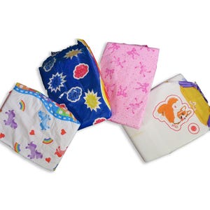 May include: Four different baby nappies with various colourful designs. One nappy has a blue background with yellow stars and white clouds, another has a pink background with pink and white figures, another has a white background with a rainbow and unicorn design, and the last one has a white background with a yellow and purple border and a cartoon dog.