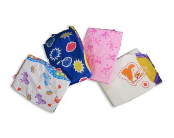 Könnte beinhalten: Vier verschiedene Babywindeln mit verschiedenen bunten Designs. Eine Windel hat einen blauen Hintergrund mit gelben Sternen und weißen Wolken, eine andere hat einen rosa Hintergrund mit rosa und weißen Figuren, eine weitere hat einen weißen Hintergrund mit einem Regenbogen und einem Einhorn-Design, und die letzte hat einen weißen Hintergrund mit einem gelben und violetten Rand und einem Cartoon-Hund.