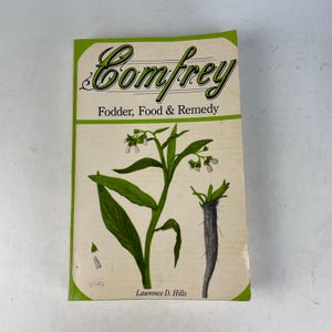 Puede incluir: Un libro antiguo titulado "Comfrey" con un borde verde. La portada presenta ilustraciones de la planta de consuelda, incluyendo hojas, flores y una raíz. El texto dice "Fodder, Food & Remedy" y el nombre del autor, Lawrence D. Hills.