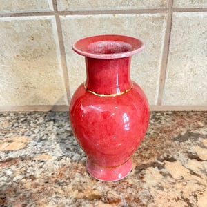 May include: A small, red vase with a glossy finish. The vase has a rounded body, a narrow neck, and a flared rim. A thin gold band encircles the neck. The vase is likely ceramic and is suitable for displaying flowers or as a decorative item.