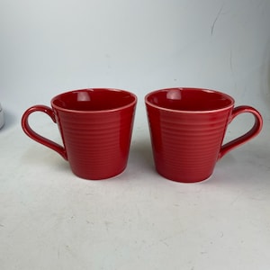 May include: Two red ceramic mugs with a ribbed texture and a white rim. Each mug has a curved handle. The mugs are set against a white background.