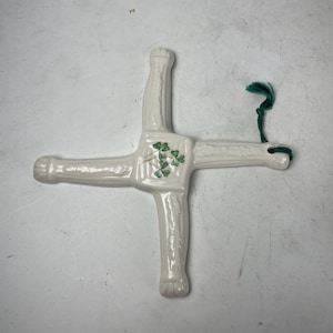 May include: A white ceramic St. Brigid's cross with a green shamrock design. The cross has four arms and a green string for hanging. The cross is a traditional Irish symbol.