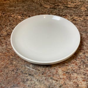 May include: A round, white ceramic plate. The plate has a slightly raised edge and a smooth, glossy surface. The plate is designed for serving food and is suitable for everyday use.