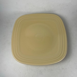 May include: A square, light yellow ceramic plate with rounded corners. The plate has concentric ridges around the edge and a circular indentation in the center. The plate is a solid color and appears to be unused.