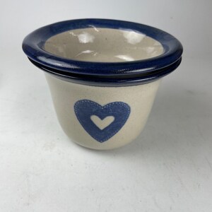May include: A ceramic planter with a cream-colored base and a dark blue rim. A blue heart is painted on the front of the planter. The planter has a wide opening and a slightly flared shape.