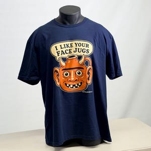 May include: A navy blue short-sleeve t-shirt featuring a graphic of an orange, horned face jug with large ears and a wide smile. Above the jug, a speech bubble contains the text I LIKE YOUR FACE JUGS in bold, dark, capitalized lettering.
