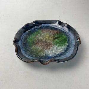 May include: A ceramic ashtray with a dark blue rim and a colorful interior. The interior features a mix of green, yellow, and brown colors, with a clear, textured surface. The ashtray has a unique, wavy edge design.