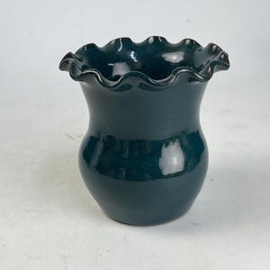 May include: A dark teal ceramic vase with a ruffled rim. The vase has a bulbous base and a flared neck. The glossy finish reflects light, highlighting the curves and the dark teal color.