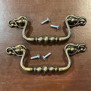 May include: Two antique brass drawer pulls with decorative scrollwork and a central beaded design. Four silver screws are included. The hardware is designed for furniture restoration or new builds.