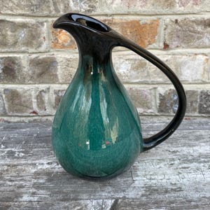 May include: A teardrop-shaped, ceramic pitcher with a glossy finish. The top and handle are black, transitioning to a deep teal green body. The handle curves gracefully from the top, creating a unique silhouette. The pitcher sits on a weathered wooden surface.