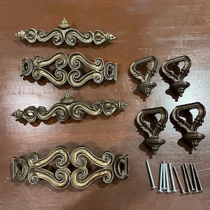 May include: A collection of antique brass cabinet hardware, including ornate pulls and handles. The set features decorative scrollwork and includes various sizes of pulls, handles, and mounting screws. The hardware is arranged on a dark wooden surface.