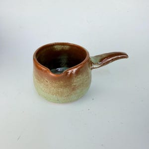 May include: A small ceramic pitcher with a round body and a single handle. The exterior features a two-tone glaze with a smooth, glossy brown finish on the upper portion and a textured, matte light green finish on the lower portion of the vessel.