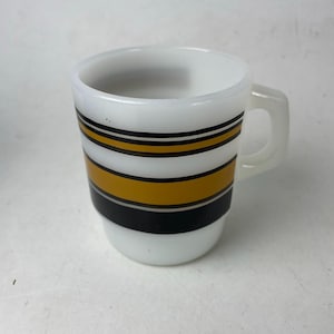 May include: White mug with a handle, featuring horizontal stripes in black and gold. The mug has a cylindrical shape and a simple, classic design. The mug is made of a glossy material.