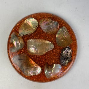 May include: Round coaster with a glittery orange resin base. The coaster is embedded with several iridescent abalone shells. The shells have a pearlescent sheen with colors of gold, silver, and green. The coaster is a decorative item.