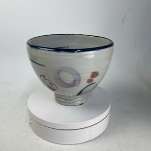 May include: A ceramic bowl with a white base, adorned with blue, purple, and red abstract designs. The rim is outlined in dark blue. The bowl is round and sits on a white surface.