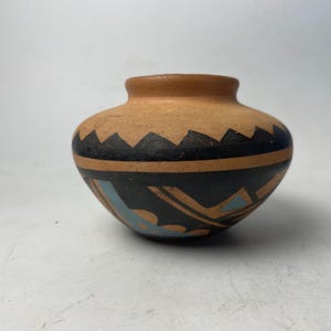 May include: A small, round, earthenware pot with a tan body and a dark rim. The pot features a black band with a zigzag pattern and blue and tan geometric designs. The pot is likely handmade and has a rustic appearance.