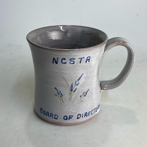 May include: A ceramic mug with a gray interior and handle, and a light gray exterior. The mug features the text "NCSTA" and "BOARD OF DIRECTORS" in blue, along with a blue floral design. The mug is suitable for hot or cold beverages.