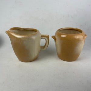 May include: A vintage ceramic creamer and sugar bowl set. The creamer has a handle and spout, while the sugar bowl has two small handles. Both pieces are a warm, two-tone color, with a light beige at the top and a darker brown at the bottom.