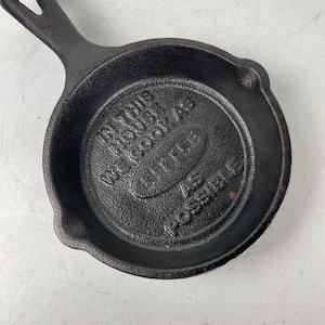 May include: A small, black cast iron skillet with a handle. The skillet has the text "IN THIS HOUSE WE COOK AS LITTLE AS POSSIBLE" embossed on the bottom. The pan is round with a small pouring spout.