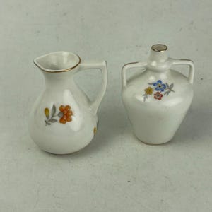 May include: Two small, white ceramic pitchers with floral designs. One pitcher has a handle and a gold rim, with orange and yellow flowers. The other has two handles and a gold rim, with blue, yellow, and red flowers.