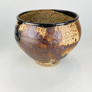May include: A rounded ceramic bowl featuring a mottled glaze in shades of dark brown, tan, and black. The exterior surface displays a textured, speckled appearance with a smooth, dark rim. The bowl sits on a small, circular base.