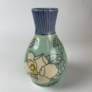 May include: A small, ceramic vase with a light blue neck and a rounded body. The body is a pale green with a large, cream-colored flower design and black leaf outlines. The flower has a yellow center.