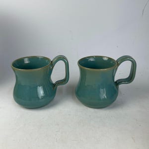 May include: Two teal ceramic mugs with curved bodies and rounded handles. The mugs have a glossy finish and a contrasting rim. The mugs are designed for beverages and are suitable for home use.