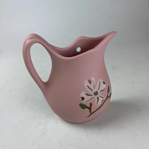 May include: A small, pink ceramic pitcher with a curved handle and a floral design. The pitcher features a white flower with green accents. The pitcher has a small hole near the top.