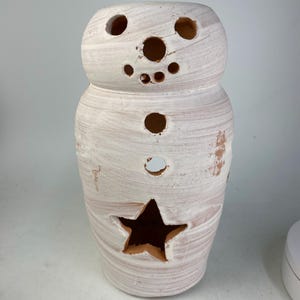 May include: A snowman-shaped candle holder with a star cutout and various circular openings. The ceramic piece is painted white with visible brushstrokes, revealing the underlying terracotta color. The design is suitable for seasonal decor.