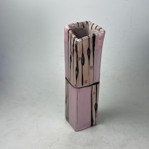 May include: A tall, rectangular ceramic vase with a pink base and a square opening. The vase features vertical black lines and tan stripes with small pink dots. A black line encircles the vase near the middle.
