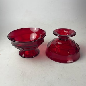 May include: Two ruby red glass dessert bowls. One bowl is upright, the other is inverted. Both bowls have a faceted design and a pedestal base. The bowls are made of a transparent, glossy glass.