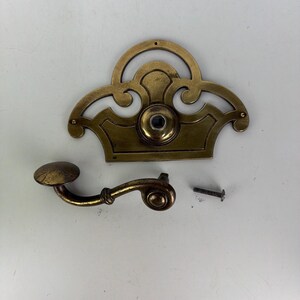 May include: A brass door handle set consisting of a decorative backplate with a central circular opening, a curved lever handle with a rounded end, and a single small metal screw. The hardware features a weathered, aged metallic finish.
