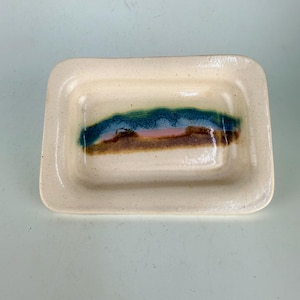 May include: A rectangular ceramic tray with a cream-colored base featuring a speckled texture. The center of the tray displays a horizontal decorative band with layered glazes in shades of deep blue, soft pink, and earthy brown.