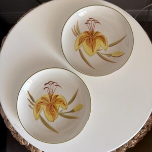 May include: Two round ceramic bowls with a cream-colored base and a green rim. Each bowl features a hand-painted yellow and brown lily flower design with green leaves. The bowls are smooth, circular, and have a shallow depth with a wide opening.