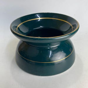 May include: A dark teal ceramic bowl with a gold trim. The bowl has a wide, shallow top and a rounded base, connected by a narrower midsection. The gold trim accents the top and base of the bowl.
