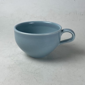 May include: A light blue ceramic teacup with a rounded bowl and a curved handle. The cup has a smooth, glossy finish and is designed for hot beverages. The teacup is a solid color and is suitable for everyday use.