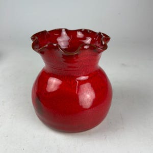 May include: A vibrant red ceramic vase with a ruffled rim and a bulbous body. The vase has a glossy finish and a dark brown trim around the rim. The vase is a decorative item.