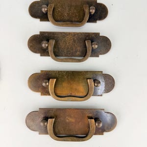 May include: A set of four rectangular brass drawer pulls with rounded ends and a weathered patina. Each pull features a central U-shaped drop handle attached to a flat backplate by two small circular studs, creating a uniform hardware set.