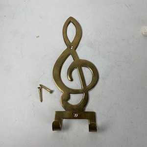 May include: A brass-colored wall hook in the shape of a treble clef. The hook has two lower prongs and includes two gold-colored screws for mounting. The design is a decorative element for music lovers.