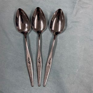 May include: Three shiny, silver spoons with oval bowls and slender handles. The handles have a textured, leaf-like design. The spoons are arranged on a light blue surface, reflecting the surrounding light.