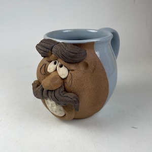 May include: A handmade ceramic mug with a whimsical face design. The mug features a brown face with a gray handle and rim. The face has dark brown mustache and eyebrows, with white eyes and a white, textured area for the mouth.
