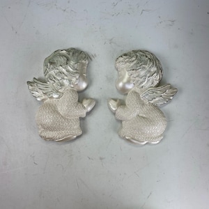 May include: Two decorative angel figurines. Each angel is in a kneeling position with hands clasped. The angels have silver wings and hair, and are wearing white robes. The figurines are made of a textured material.
