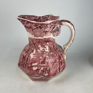 May include: A vintage ceramic pitcher with a cream-colored handle and rim. The pitcher features a detailed red and white transferware design depicting a landscape scene with figures and buildings. The pitcher has a unique, flared spout and a bulbous body.
